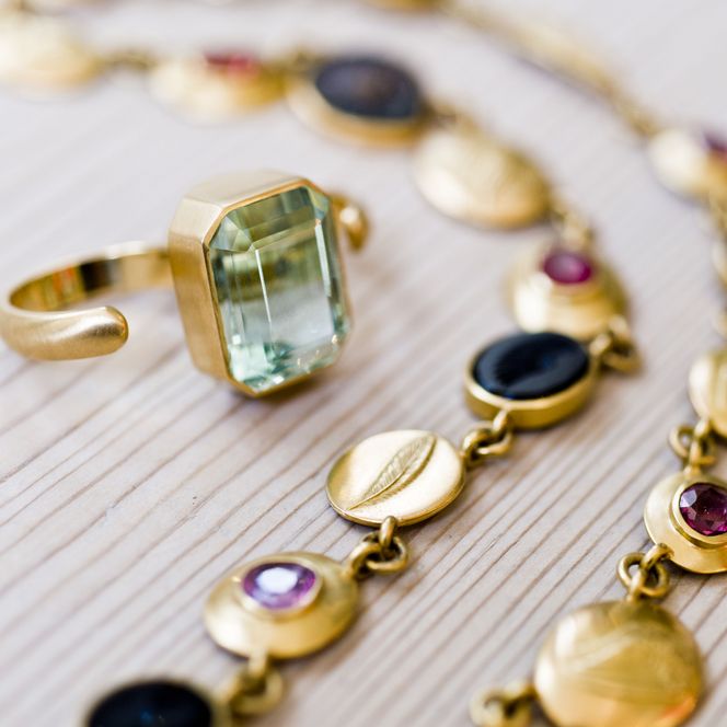 Goldschmuck von Goldschmied Christoph Freier Goldener Ring mit grünem Stein und farbiger Kette auf Holzuntergrund.