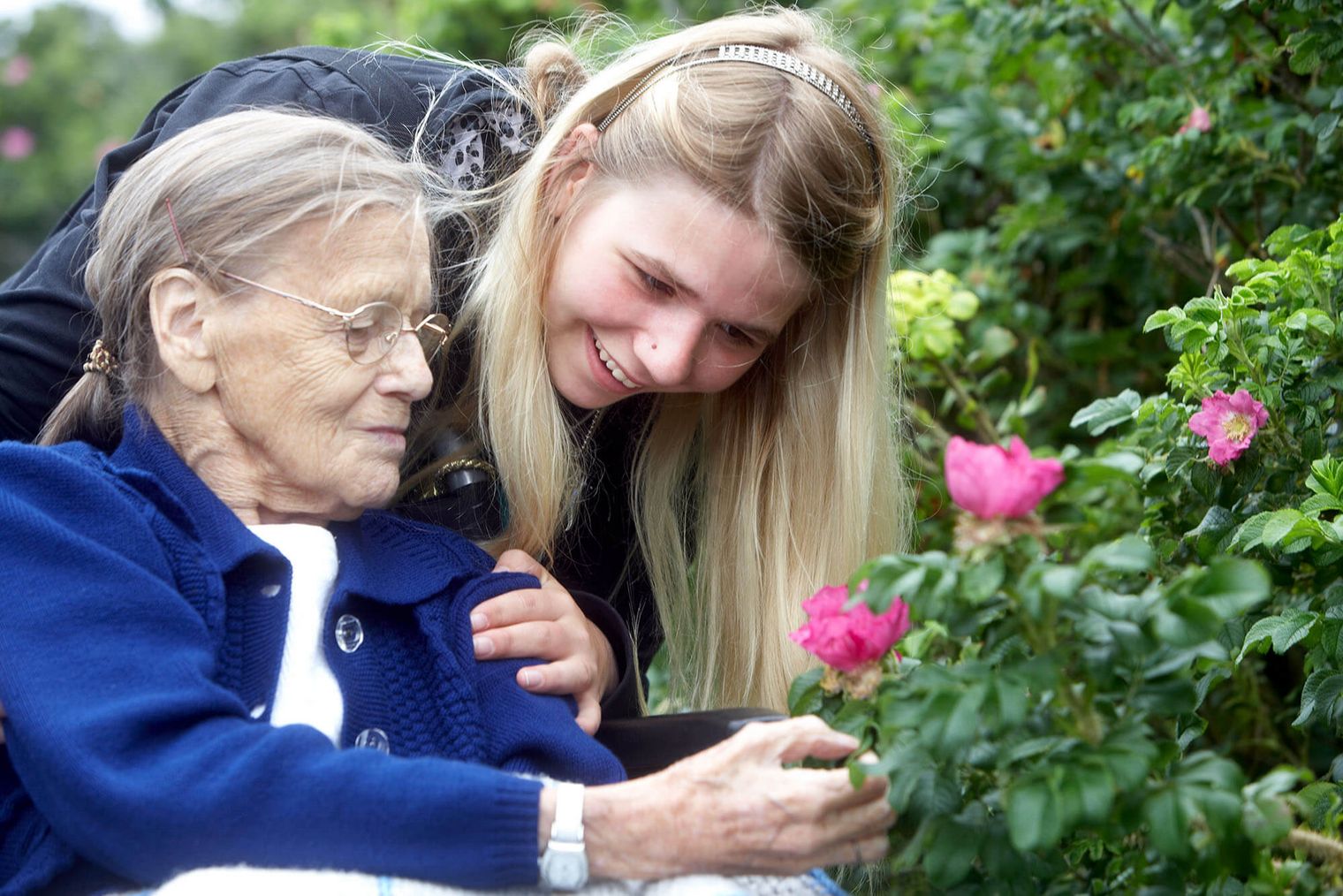 Eine junge Frau beugt sich lächelnd zu einer älteren Dame, die eine rosa Blüte betrachtet. Beide sind von grünen Pflanzen und Blüten umgeben.