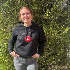 Eine junge Frau mit dunklem Haar steht draußen vor einem grünen, dicht bewachsenen Busch. Sie trägt einen schwarzen Kapuzenpullover mit dem Logo der Azubi-Crew Sylt 2024 und hat ihre Hände in den Hosentaschen. Sie lächelt freundlich in die Kamera.