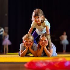 Drei Kinder spielen auf einer Bühne; ein Mädchen balanciert auf den Schultern von zwei sitzenden Kindern.
