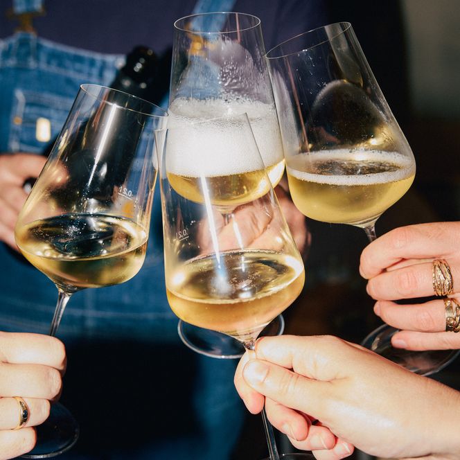 Vier Gläser mit Weißwein stoßen an, gesellige Stimmung, Person im Hintergrund unscharf sichtbar.