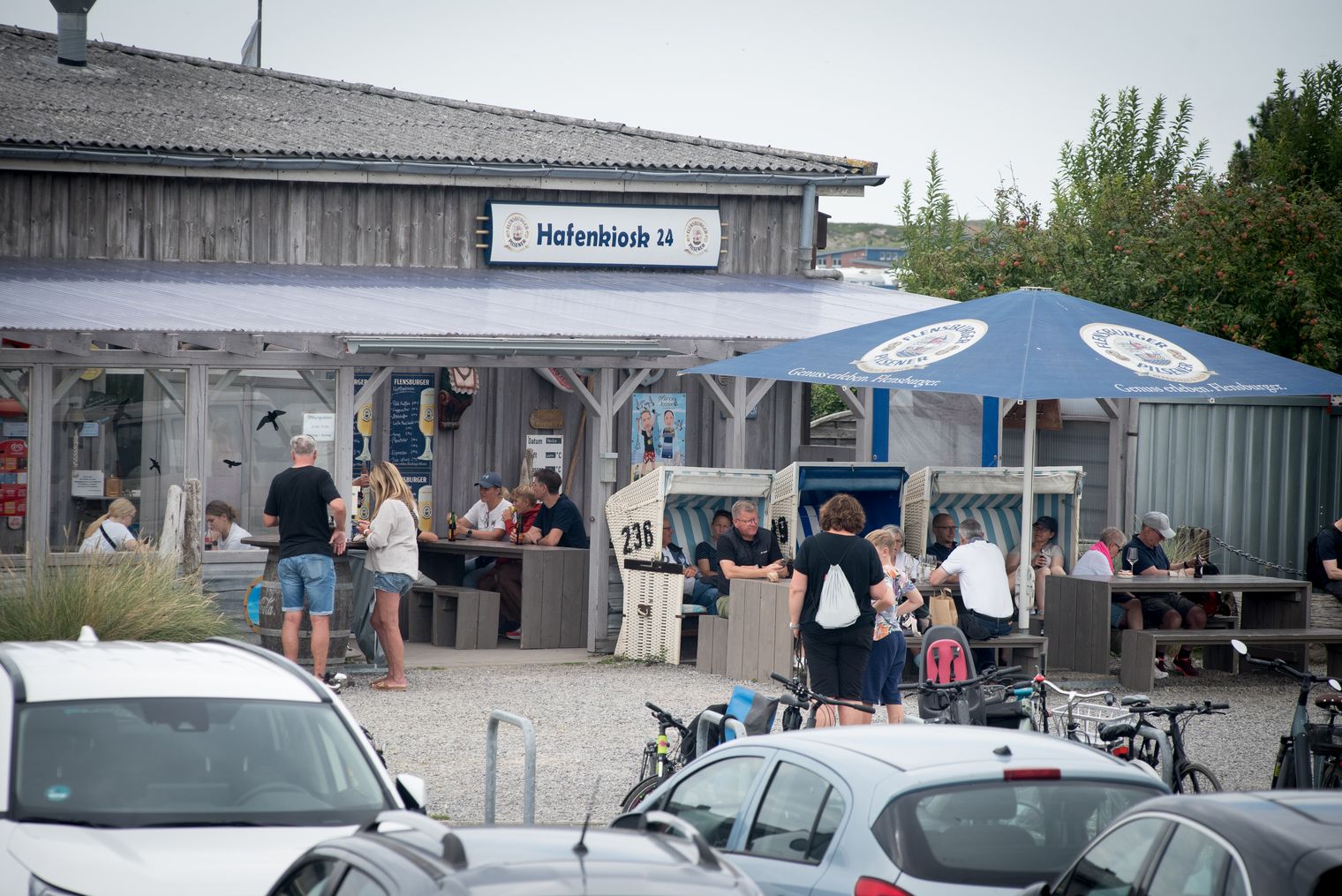 Menschen sitzen und stehen vor dem Hafenkiosk 24 mit Strandkörben und Sonnenschirm.