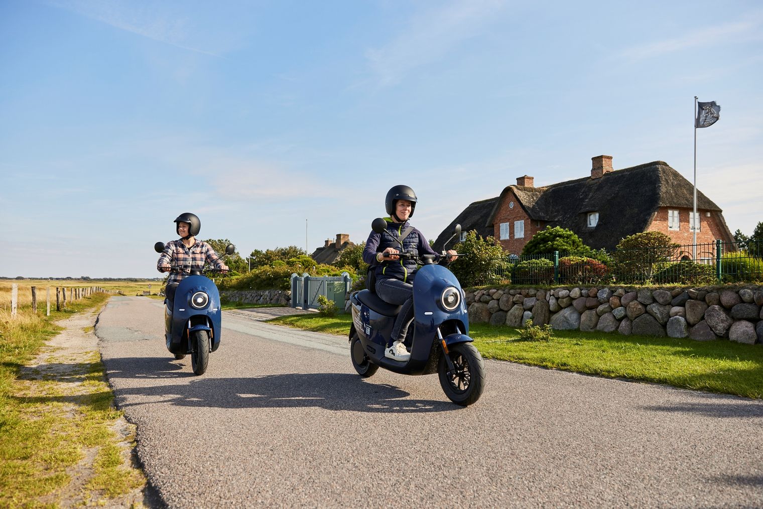 Zwei Menschen fahren auf blauen E-Rollern eine Landstraße entlang, vorbei an einem Reetdachhaus auf Sylt.