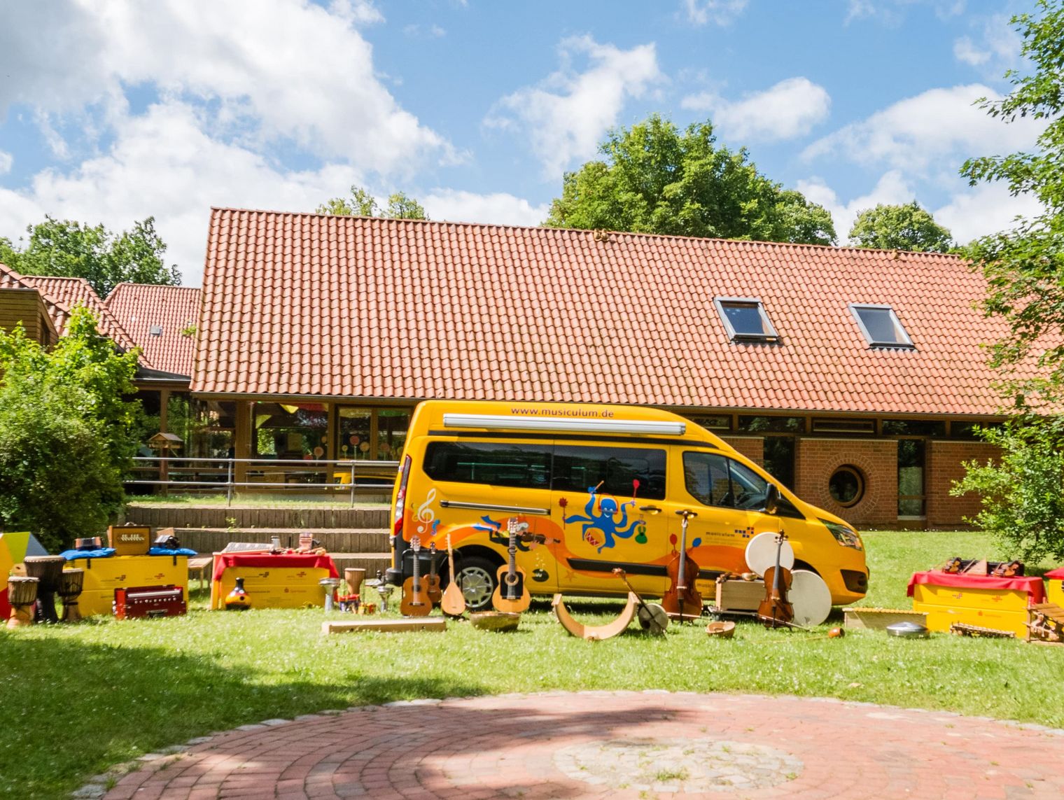 Gelber Musikbus mit Instrumenten im Freien vor einem Gebäude mit rotem Dach.