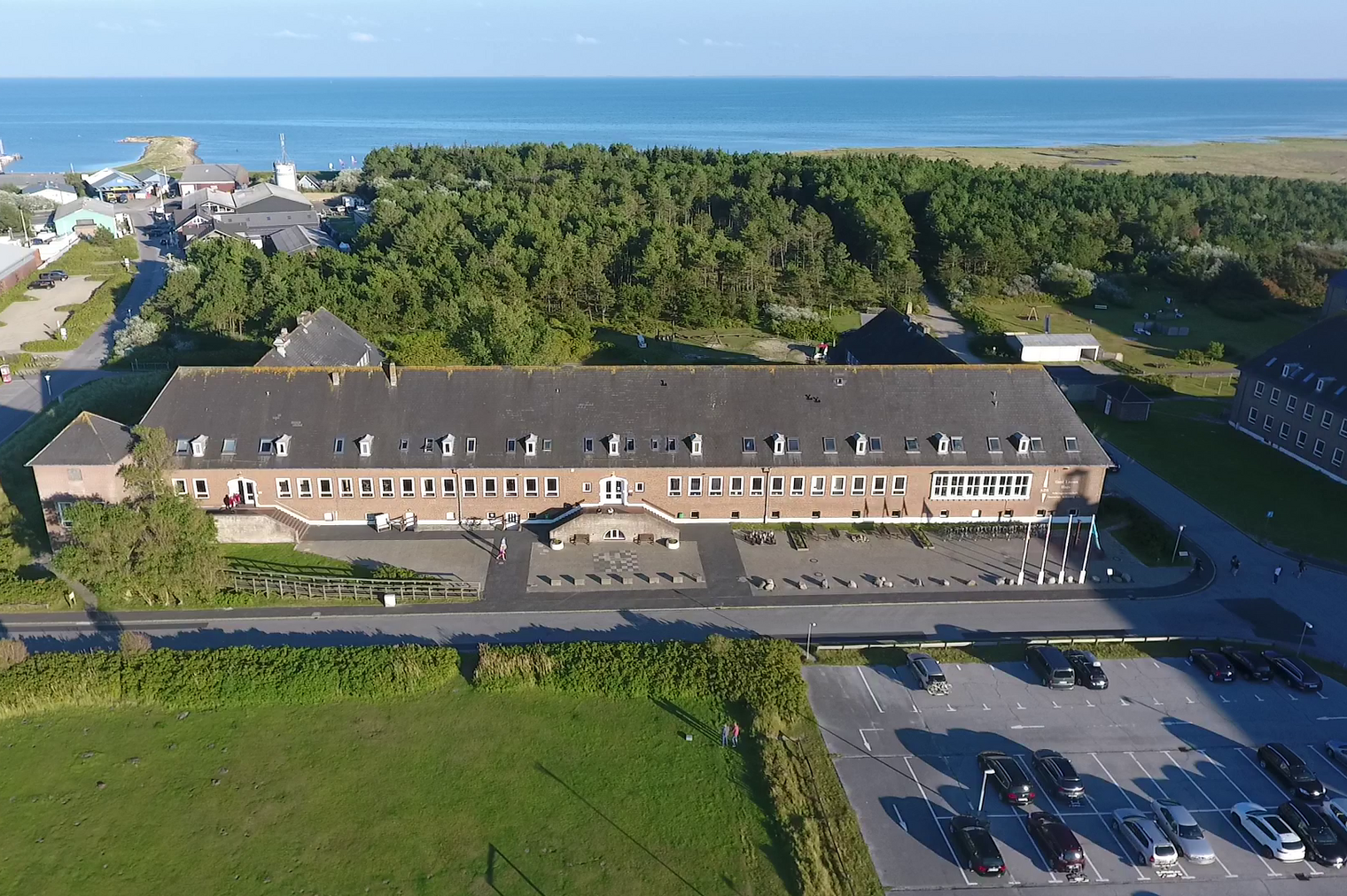 Großes Backsteingebäude vor Wald, Dünen und Nordsee, mit Parkplatz im Vordergrund.
