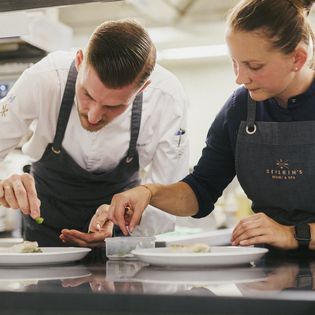 Zwei Köche, ein Mann und eine Frau, richten Speisen auf weißen Tellern in der Küche des Severin's Resort & Spa in Keitum auf Sylt an.