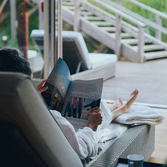 Entspannung pur im Severin*s Resort & Spa Eine Person in einem weißen Bademantel liest auf einer Liege ein Magazin, im Hintergrund ist eine Holzterrasse und grüne Vegetation sichtbar.