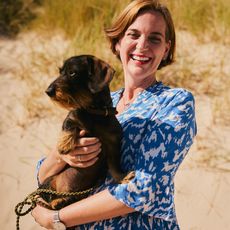 Frau in einem blauen gemusterten Kleid hält einen Dackel und steht vor einer Dünenlandschaft, beide wirken entspannt und glücklich. 