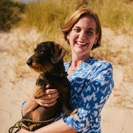 Frau in einem blauen gemusterten Kleid hält einen Dackel und steht vor einer Dünenlandschaft, beide wirken entspannt und glücklich. 