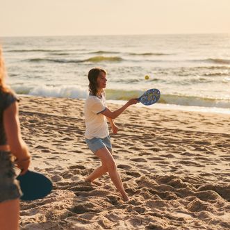 Drei Jugendliche spielen mit Schlägern und einem Ball am sonnigen Sandstrand vor dem Meer.