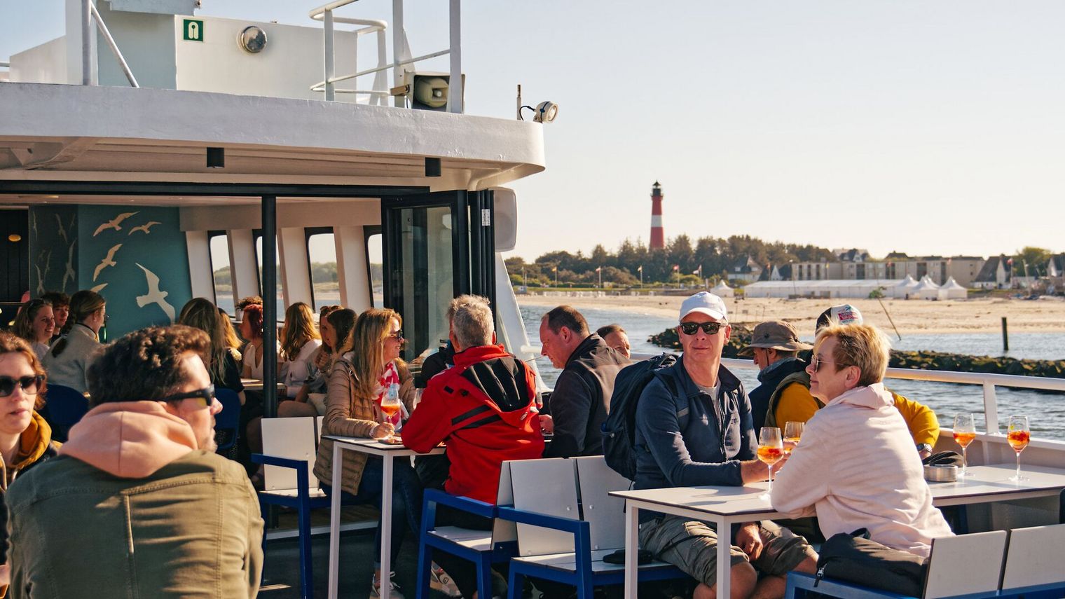 Gäste sitzen an Deck eines Schiffs mit Leuchtturm im Hintergrund