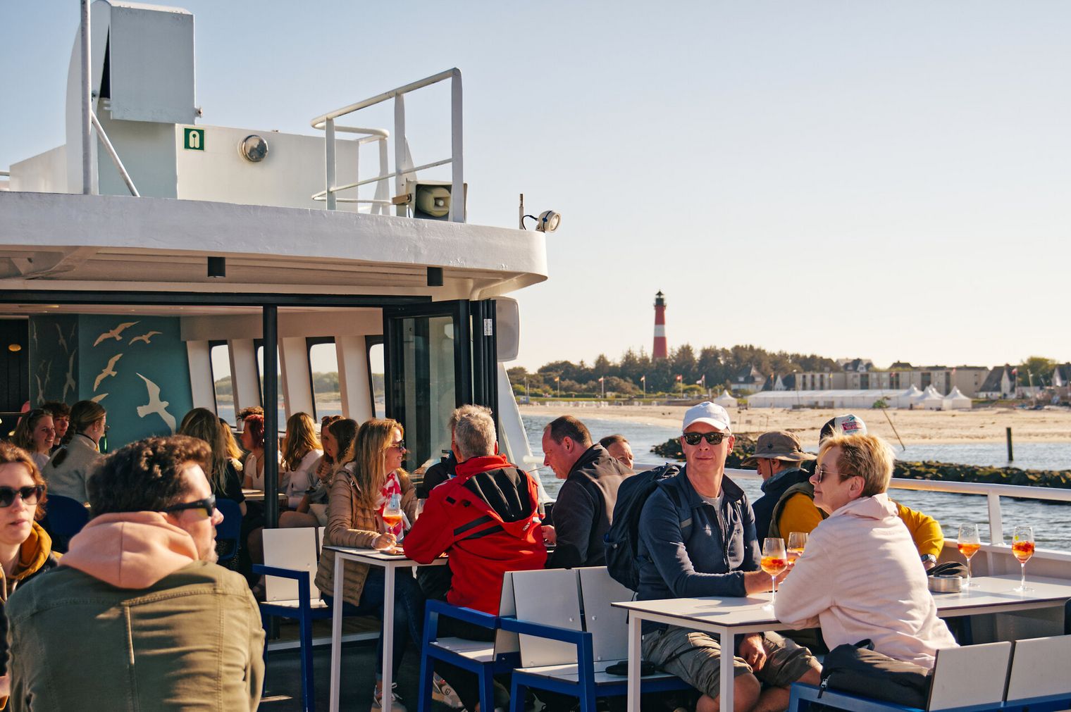 Gäste sitzen an Deck eines Schiffs mit Leuchtturm im Hintergrund