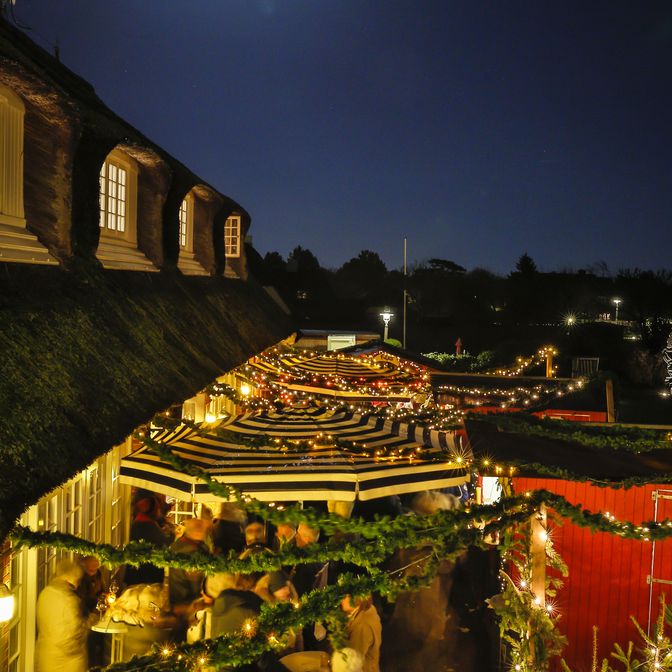 Kampen-Axel-Steinbach-Weihnachtsmarkt Weihnachtsmarkt in Kampen bei Nacht, beleuchtet von Lichterketten und einem strahlenden Vollmond, mit geschmückten Marktständen und Reetdachhäusern.
