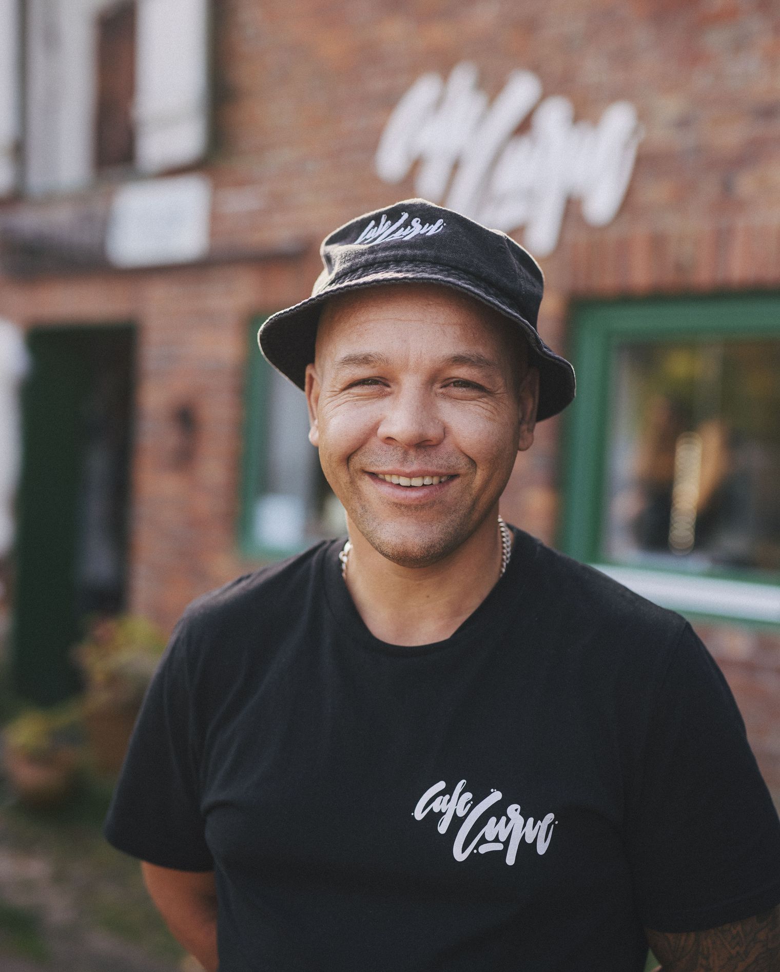 Ein Mann in schwarzem T-Shirt und Fischerhut mit Café Curve-Logo lächelt in die Kamera vor einem Backsteingebäude.