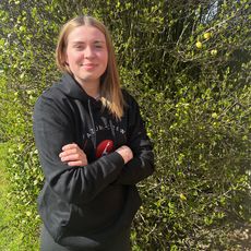 Eine junge Frau mit blonden Haaren steht draußen vor einem dichten, grünen Busch mit kleinen Knospen. Sie trägt einen schwarzen Kapuzenpullover mit der Aufschrift „Azubi-Crew“ und hat die Arme verschränkt. Sie lächelt freundlich in die Kamera.
