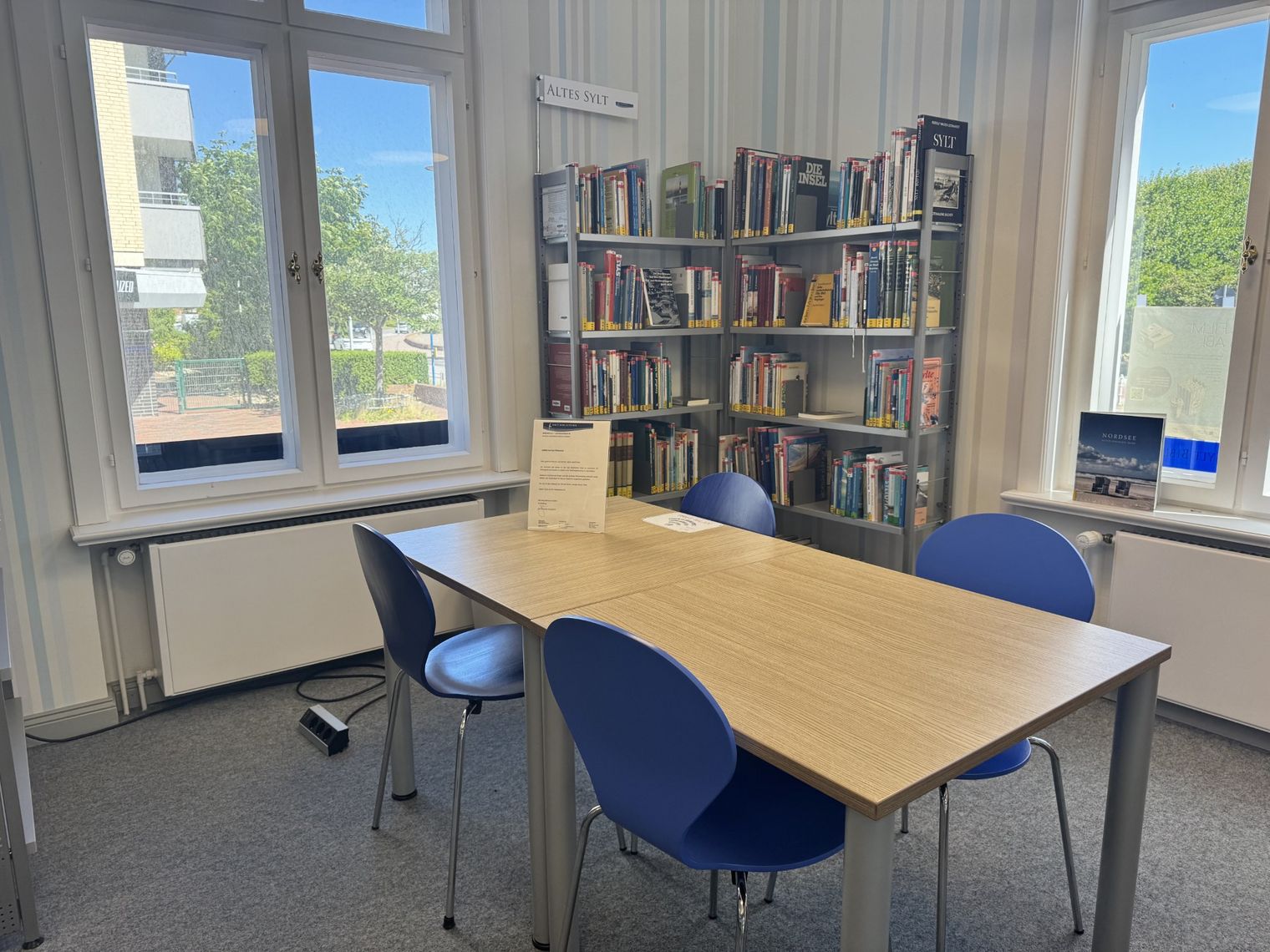 Lesetisch mit vier blauen Stühlen vor Bücherregalen und großen Fenstern in einer Bibliothek auf Sylt