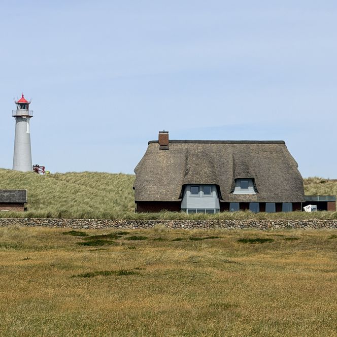 Du siehst den Leuchtturm List-Ost mit rotem Dach hinter einer Düne. Davor steht ein Haus mit Reetdach, eine Steinmauer und trockenes Gras unter blauem Himmel.