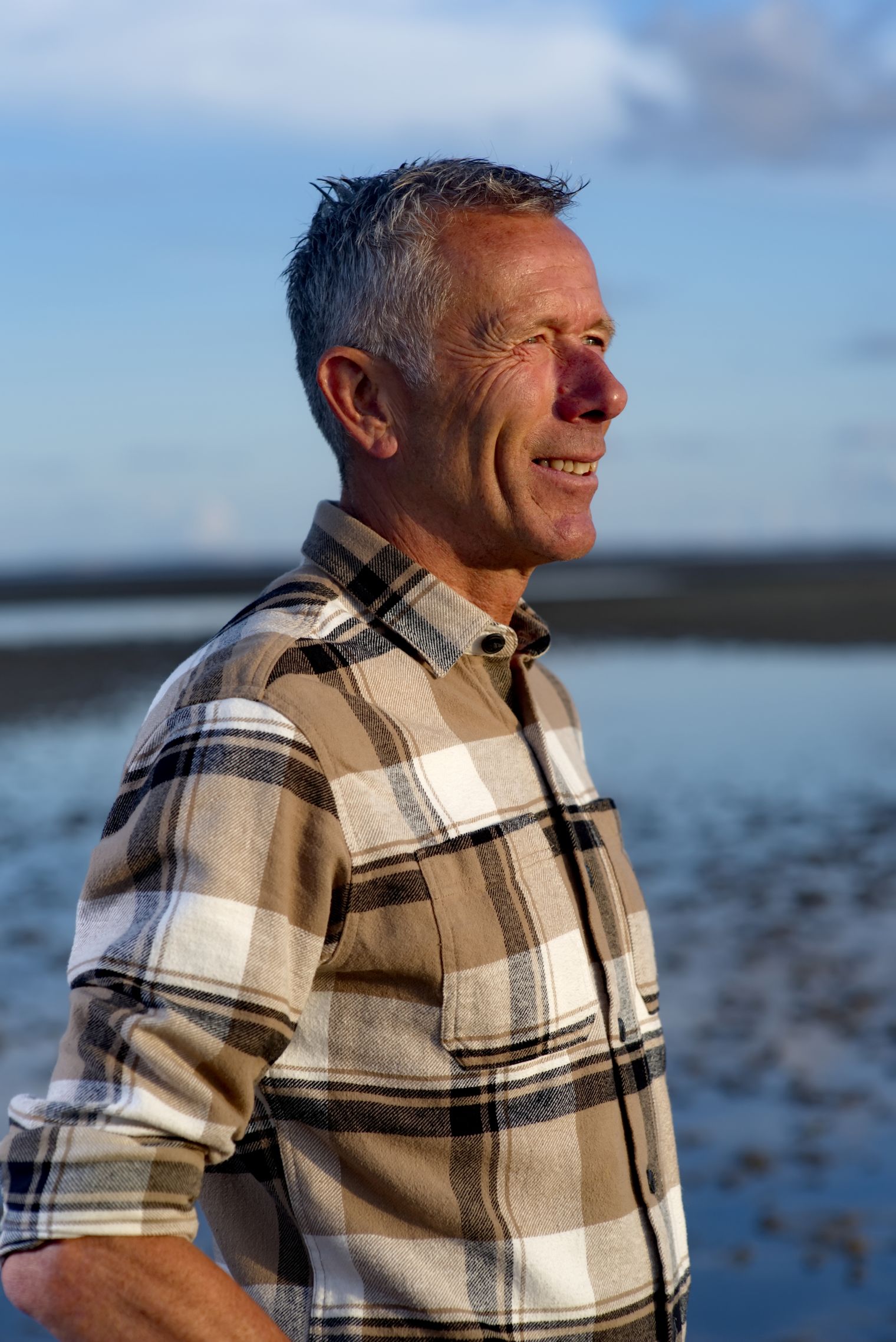 Mann im karierten Hemd steht lächelnd vor dem Wattenmeer im warmen Abendlicht.