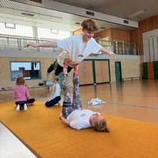 Ein Kind balanciert mit ausgebreiteten Armen auf den Füßen eines anderen Kindes in einer Turnhalle.