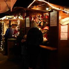 Besucher genießen einen festlich beleuchteten Weihnachtsmarktstand mit Glühwein und Flammkuchen.