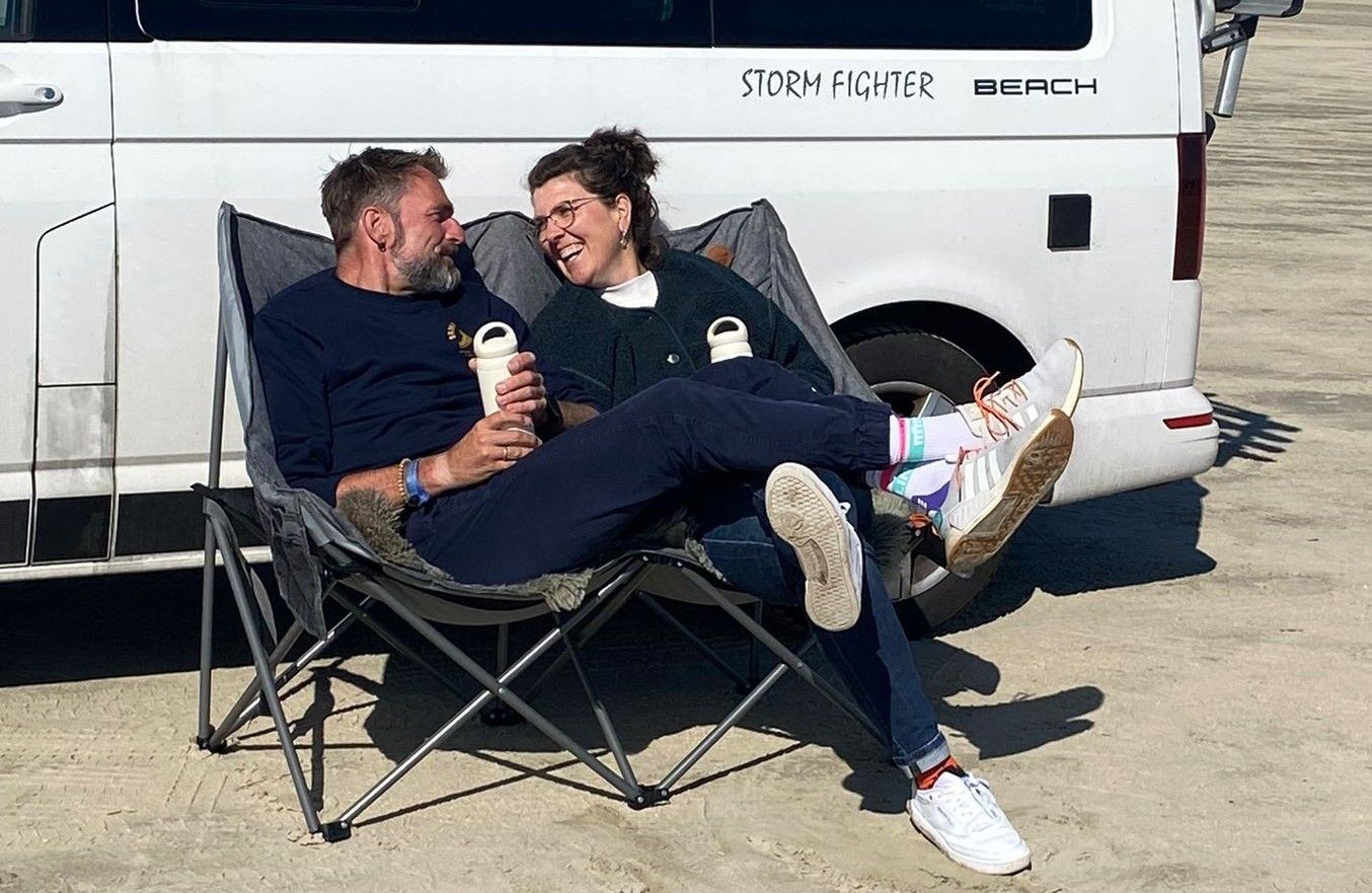Ein Paar sitzt fröhlich lachend auf Campingstühlen vor einem Bus an einem Strand.