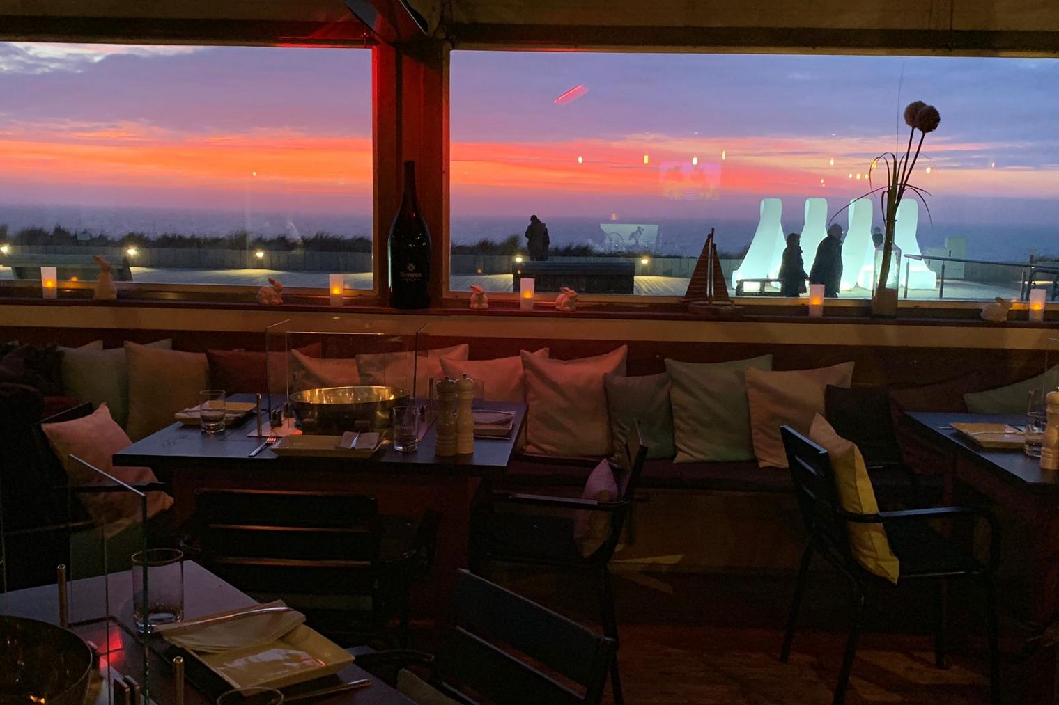 Restaurant „iismeer“ auf Sylt mit gedeckten Tischen und Blick auf einen Sonnenuntergang über den Dünen. Draußen leuchtende Skulpturen.
