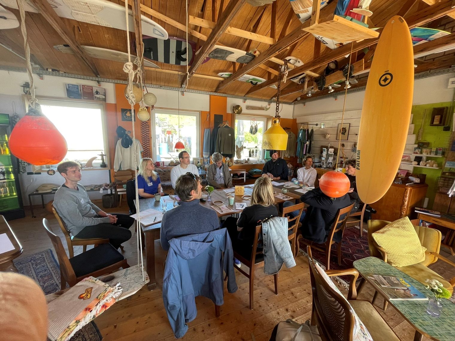 Gruppe von Menschen bei einem Treffen an einem Tisch in einem Surfshop mit hängenden Surfbrettern.
