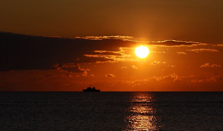 Ein Schiff fährt vor einem leuchtenden Sonnenuntergang auf dem Meer.