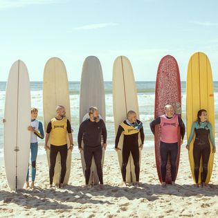 Neun Surfer stehen am Strand mit aufgestellten Longboards.