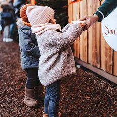 Zwei Kinder auf einem Weihnachtsmarkt, in warme Winterkleidung gehüllt, erhalten eine warme Speise oder ein Getränk an einem Marktstand.