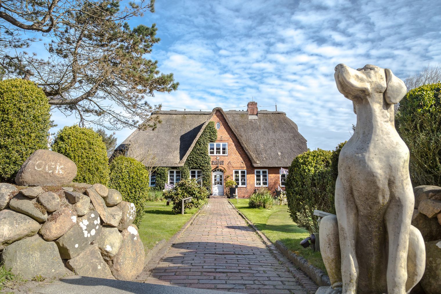 Reetgedecktes Backsteinhaus mit gepflegtem Garten, gepflastertem Weg und großer Steinskulptur im Vordergrund.