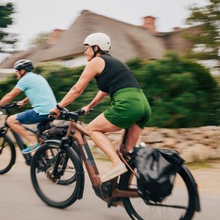 Zwei Personen auf E-Bikes fahren durch Keitum auf Sylt. Im Hintergrund sind Reetdachhäuser sichtbar.