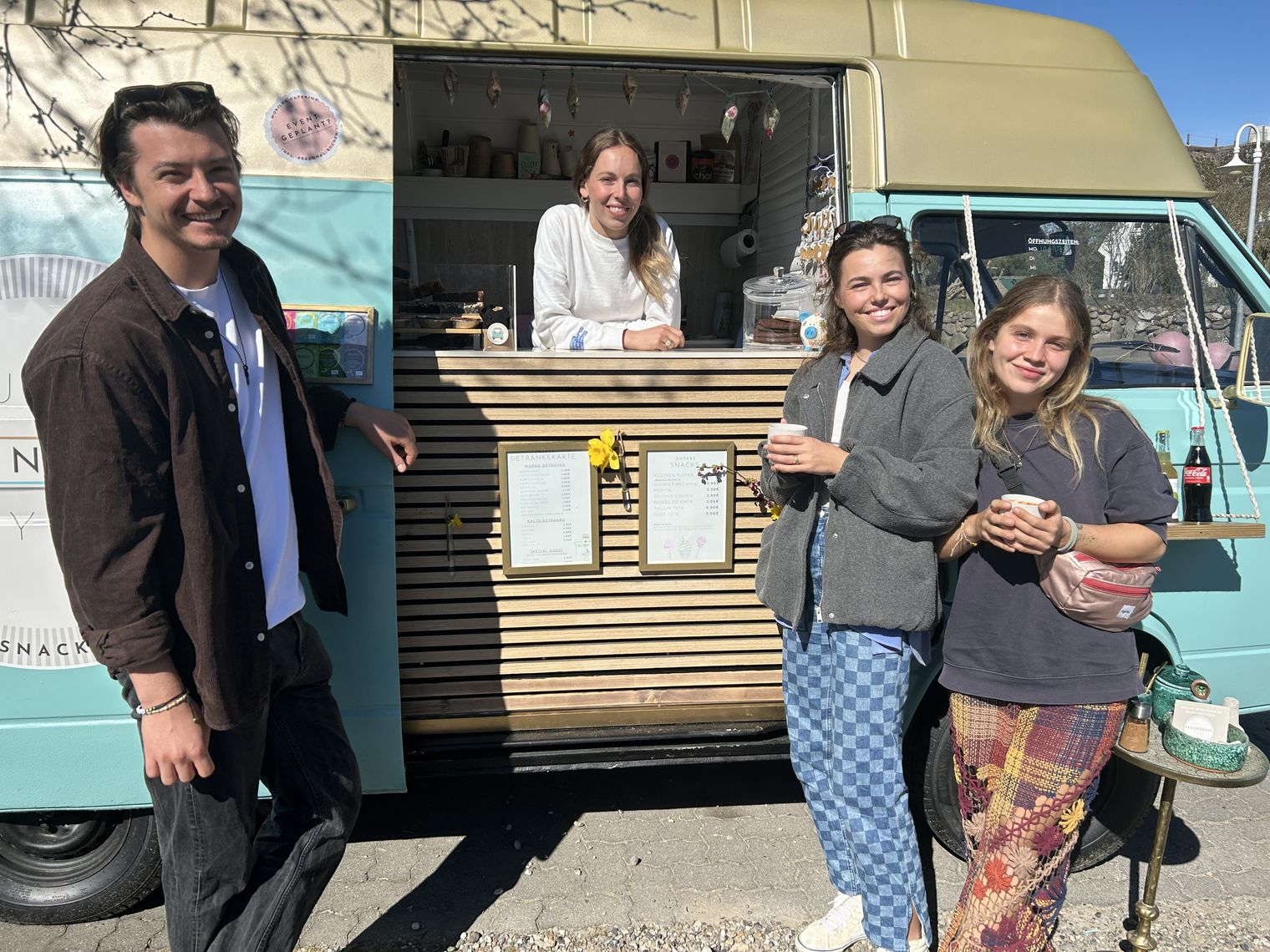Drei junge Menschen und eine Verkäuferin am Bulli Küstebohne genießen Kaffee in der Sonne.