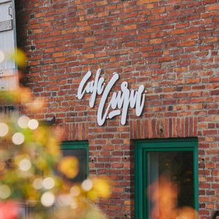 Ceglany dom z białym napisem „Café Curvy” i dwoma zielonymi oknami, na pierwszym planie rozmyte rośliny.