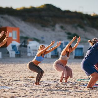 Mehrere Personen, darunter Schwangere und Senioren, üben eine Hockpose auf Handtüchern am Strand.