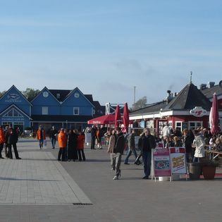Menschen am Lister Hafen mit Restaurants, blauen Gebäuden und Sitzgelegenheiten unter roten Sonnenschirmen.