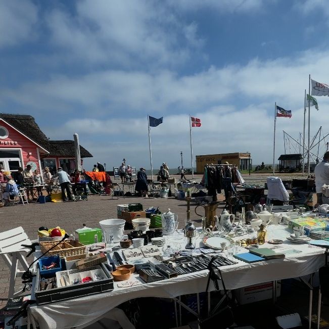 Flohmarktstand am Hafen von List auf Sylt mit Geschirr, Besteck, Kleidung und weiteren gebrauchten Gegenständen vor blauem Himmel und dem Meer.