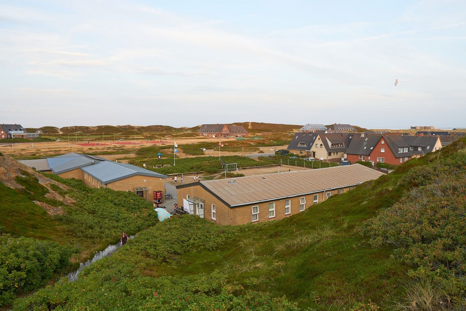 Gebäudekomplex inmitten von Dünen, umgeben von Natur und angrenzenden Ferienhäusern.