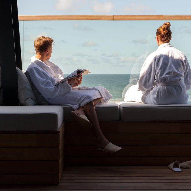 wyn-strandhotel-wellness Zwei Personen in Bademänteln entspannen auf einer Terrasse mit Meerblick und blauem Himmel.