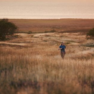 Eine Frau joggt durch hohe Gräser in der Morgendämmerung, mit Meer und Heidelandschaft im Hintergrund.