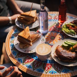 Bunter Mosaiktisch mit Sandwiches, Croissants, Kaffee und Getränken zum Frühstück.