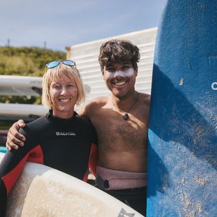 Zwei Surfer mit blauem und weißem Surfboard lächeln vor einer Surfschule.