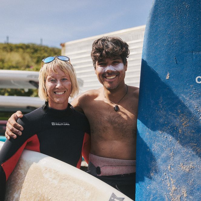 Zwei Surfer mit blauem und weißem Surfboard lächeln vor einer Surfschule.