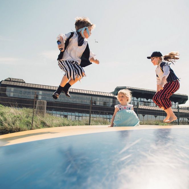 Drei Kinder spielen auf einer Hüpfburg im Freien. Zwei sind als Piraten verkleidet, mit gestreiften Hosen und Westen, während das dritte Kind ein Sommerkleid trägt. Im Hintergrund sind grüne Dünen und ein Gebäude mit großen Fenstern zu sehen.