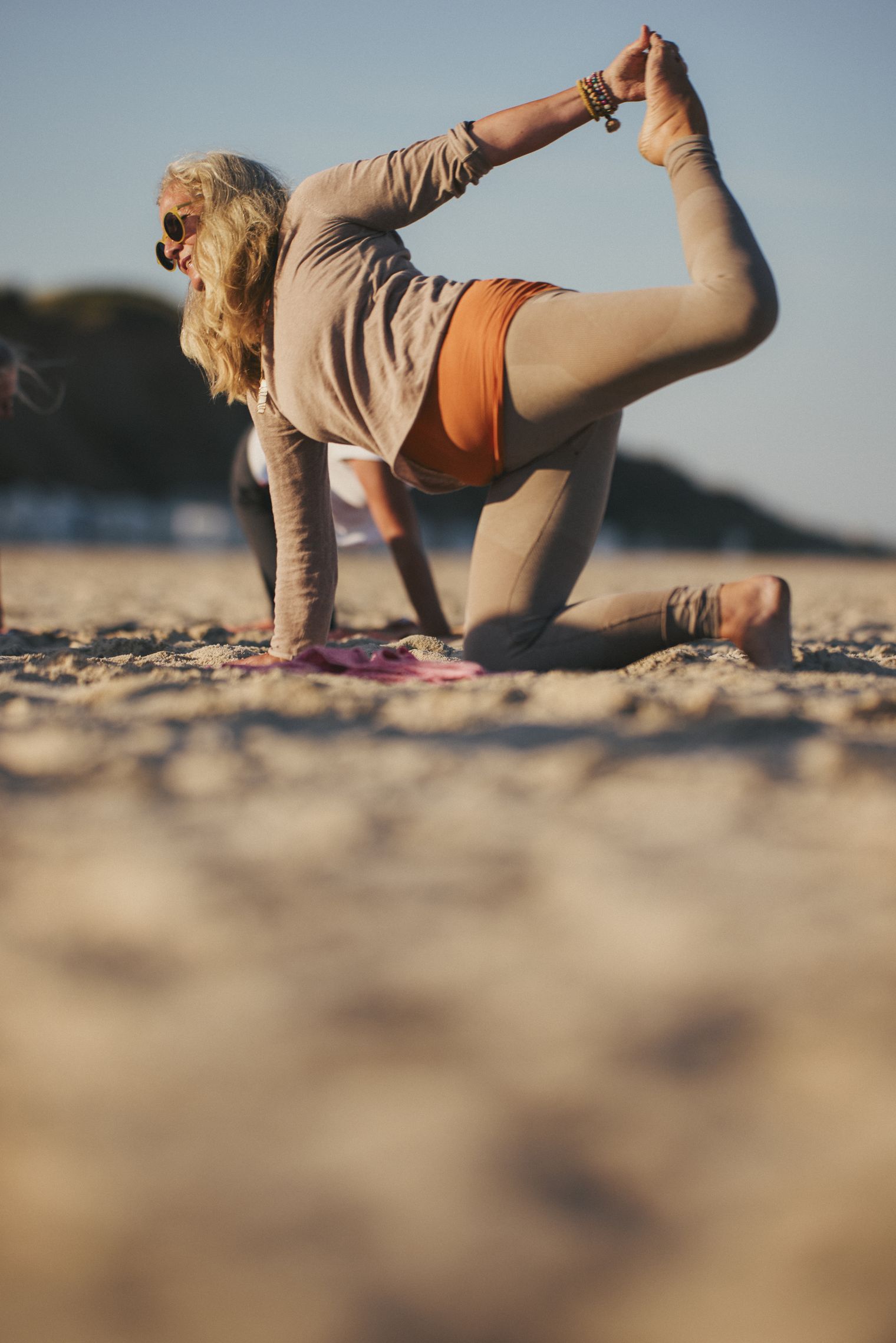 Frau beim Yoga am Strand, eine Hand hält ihren Fuß in einer gestreckten Position, andere Teilnehmer im Hintergrund. 