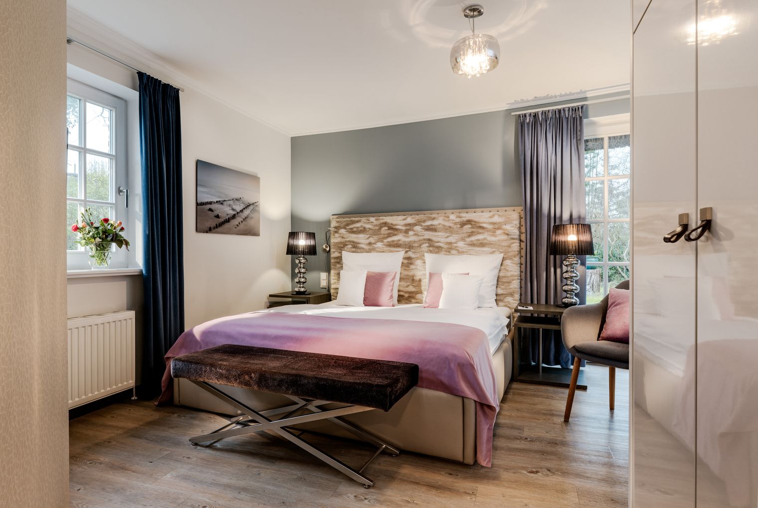 Helles Schlafzimmer mit Doppelbett, gepolstertem Kopfteil, rosa Tagesdecke, großen Fenstern und dunklen Vorhängen.