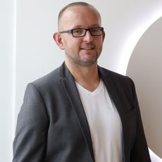 Mann mit Brille, dunklem Sakko und weißem T-Shirt lächelt vor heller Wand mit Lichtbogen.