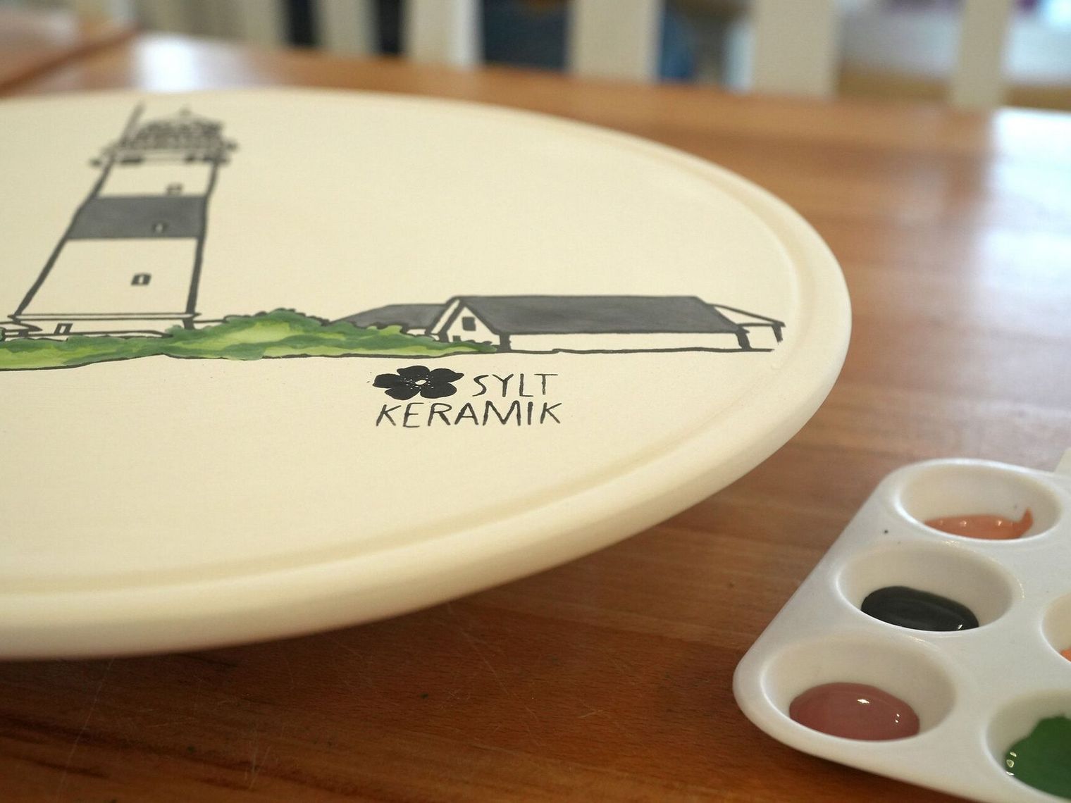 Kreative Auszeit mit Sylt-Keramik Keramikteller mit Leuchtturmmotiv und Aufschrift „Sylt Keramik“, daneben eine Farbpalette mit mehreren Farbtupfern.