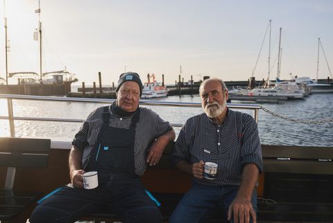 Zwei ältere Männer in gestreiften Hemden sitzen entspannt auf einem Boot im Hafen und halten Kaffeetassen in den Händen.