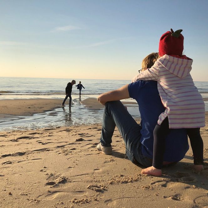 Ein Mann sitzt mit einem Kind auf dem Schoß am Strand, zwei weitere Kinder spielen am Wasser, Sonnenuntergang im Hintergrund.